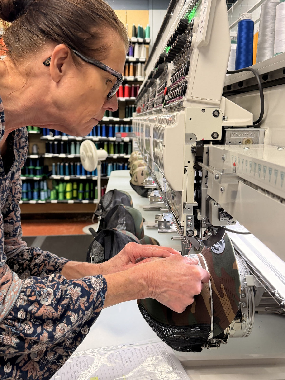 Team member hooping a camo hat on a Tajima multi-head embroidery machine, with walls of thread cones behind her