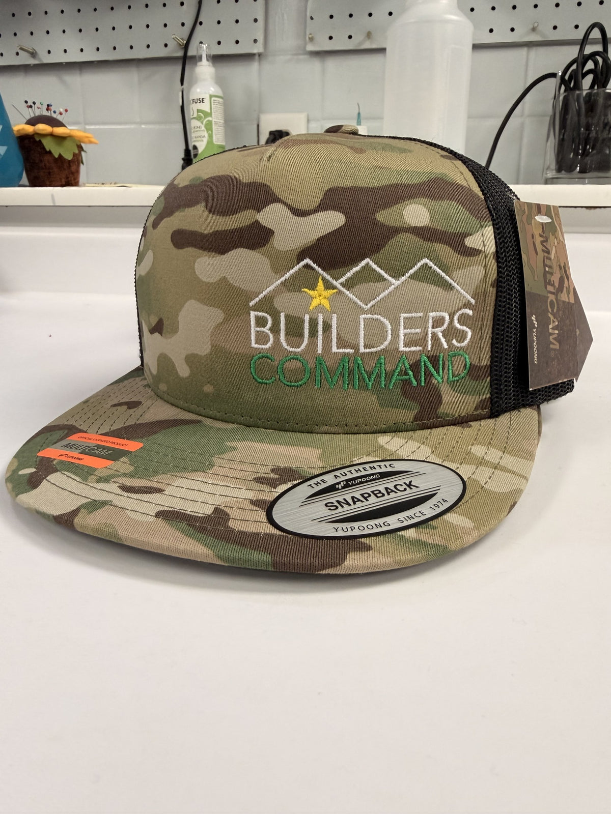 Multicam snapback trucker hat embroidered with the Builders Command logo: a white mountain roofline with a gold star, BUILDERS in white and COMMAND in green