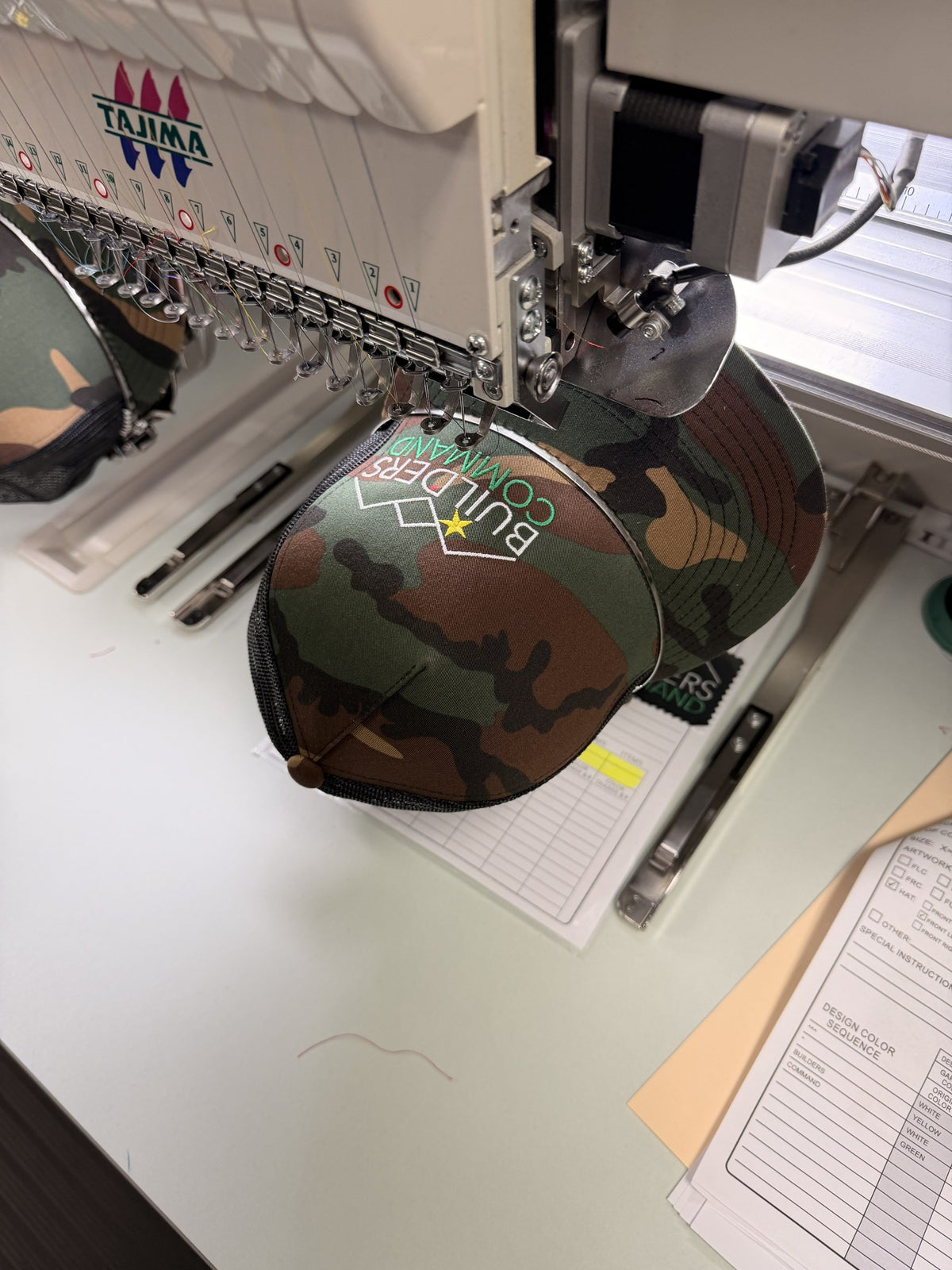 Camo hat on a Tajima embroidery machine with the Builders Command logo freshly stitched in white, green and gold