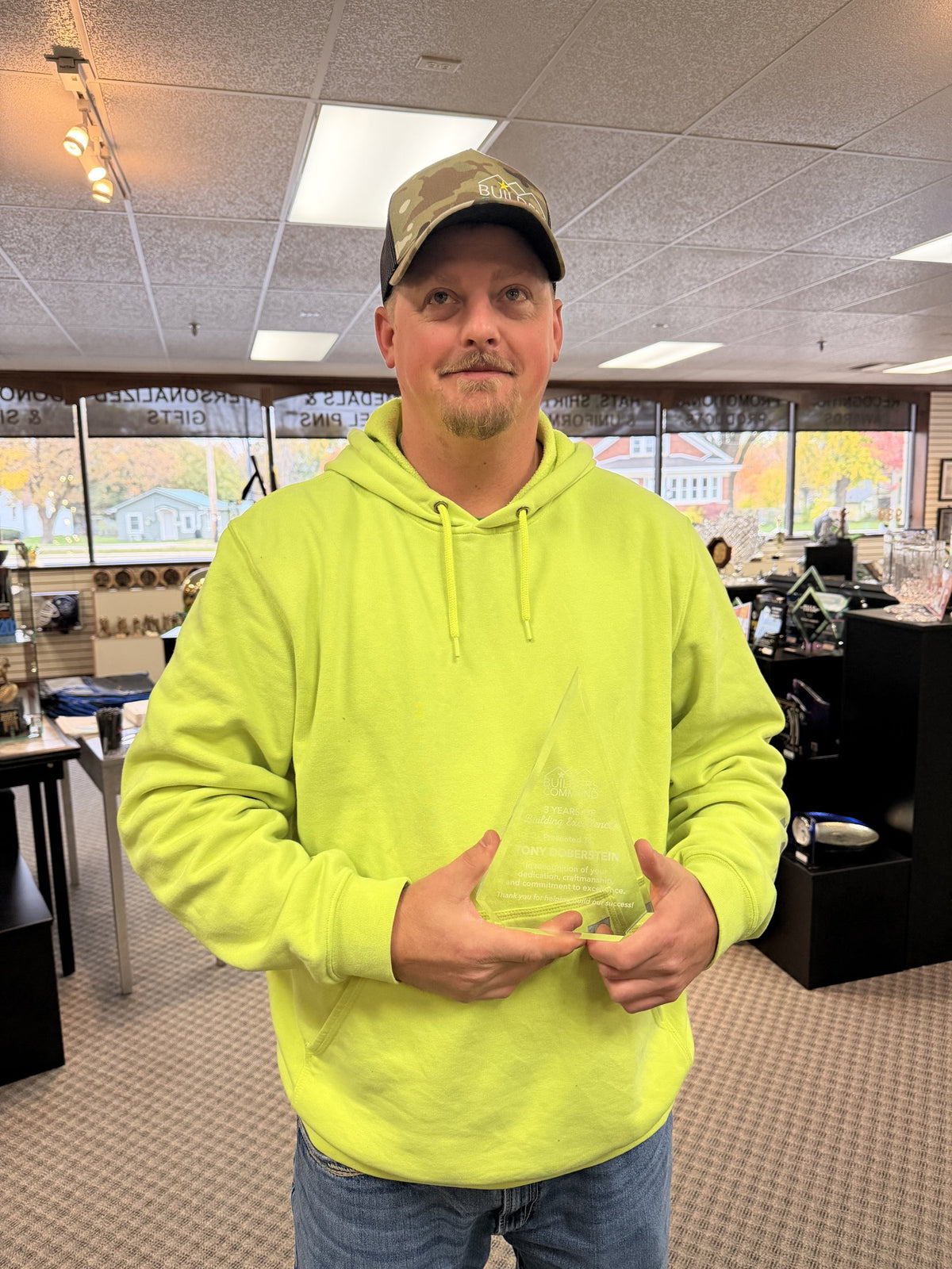 Josh, owner of Builders Command, holding one of the clear acrylic awards in our showroom, wearing a camo hat embroidered with the Builders Command logo